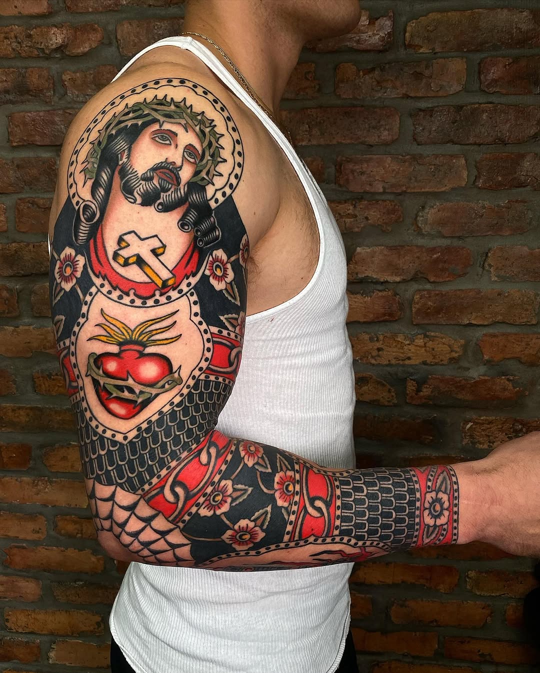 American traditional sleeve featuring a sacred heart Christ figure with halo, fish scales, and decorative floral elements in red and black.