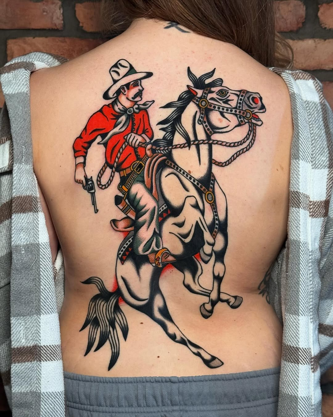 Cowboy on rearing horse with decorative saddle and tack, rendered in bold lines with red shirt and natural skin tones, spanning upper back.
