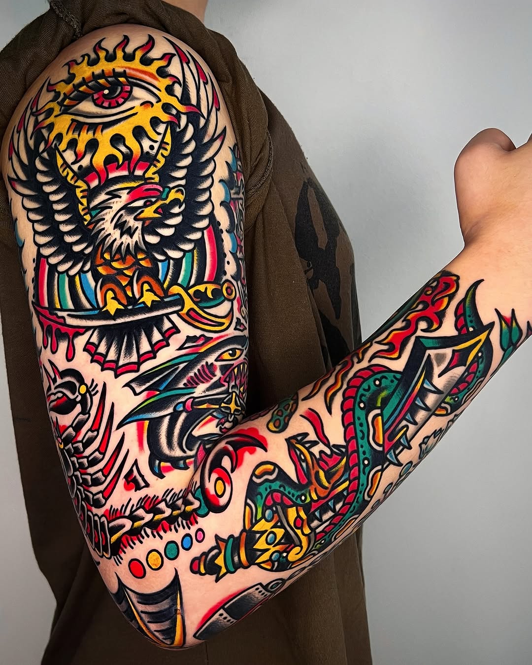 Full sleeve American traditional tattoo featuring eagle, skull, fish, and ornamental designs in bold black outlines with saturated primary colors.