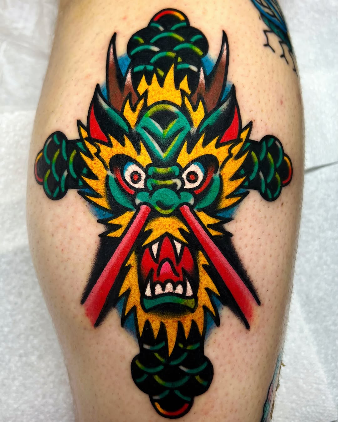 Symmetrical demon or oni face with yellow spikes, green scales, red eyes, and red laser beams on calf.