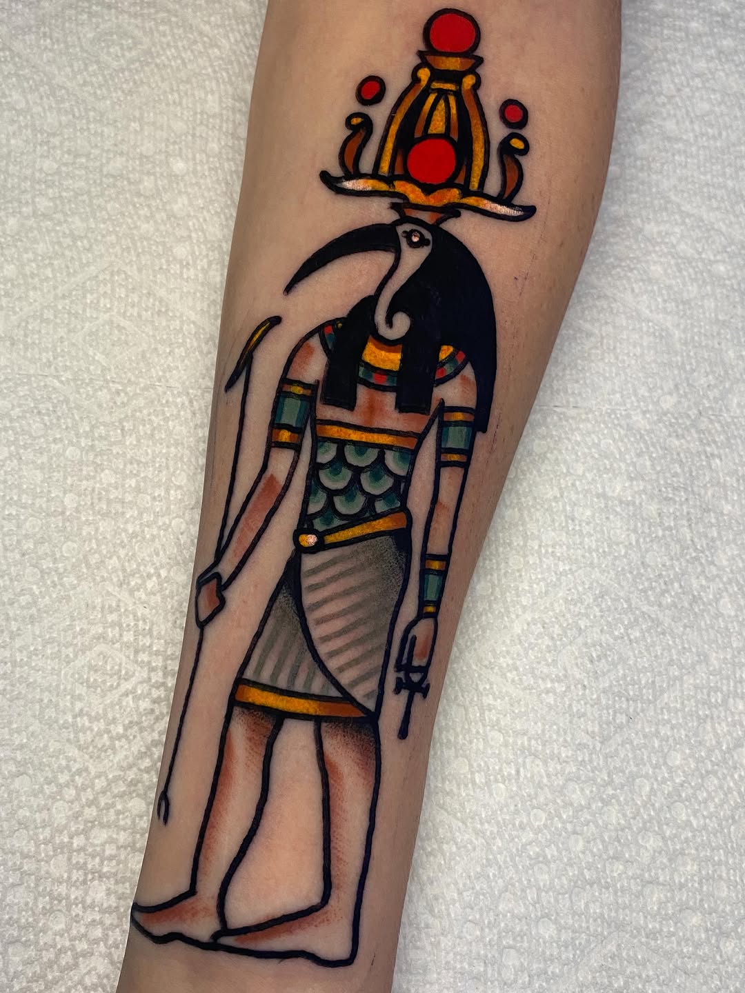 Egyptian figure with ibis head holding a ceremonial vessel, rendered in bold outlines with traditional saturated colors on forearm.