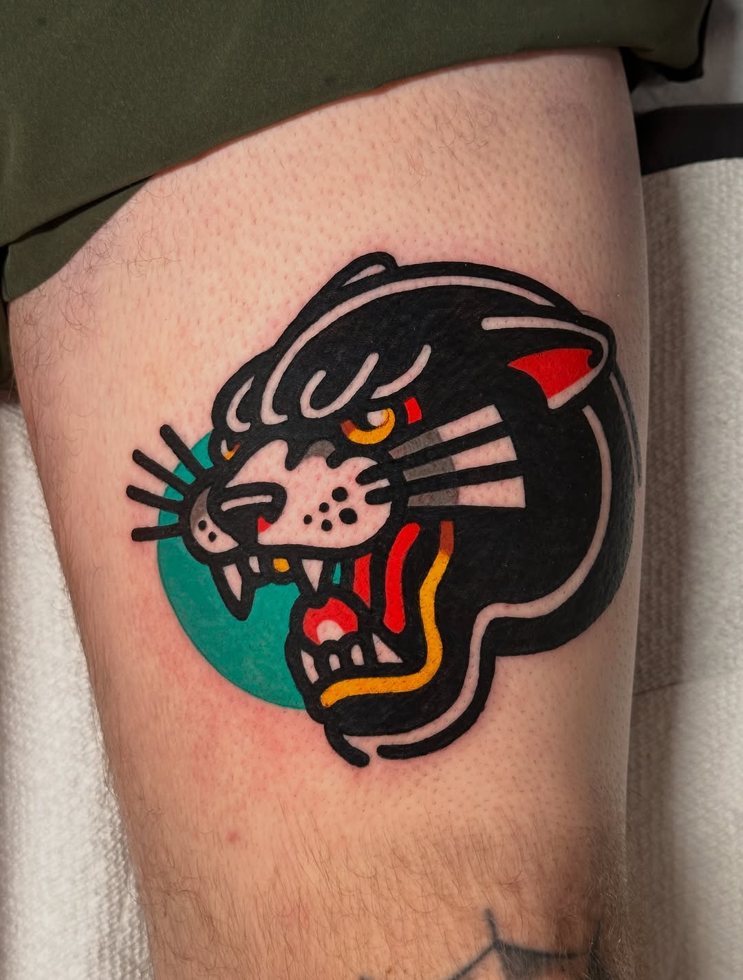 Black panther head with red inner ear and golden eye over green circular background on thigh.