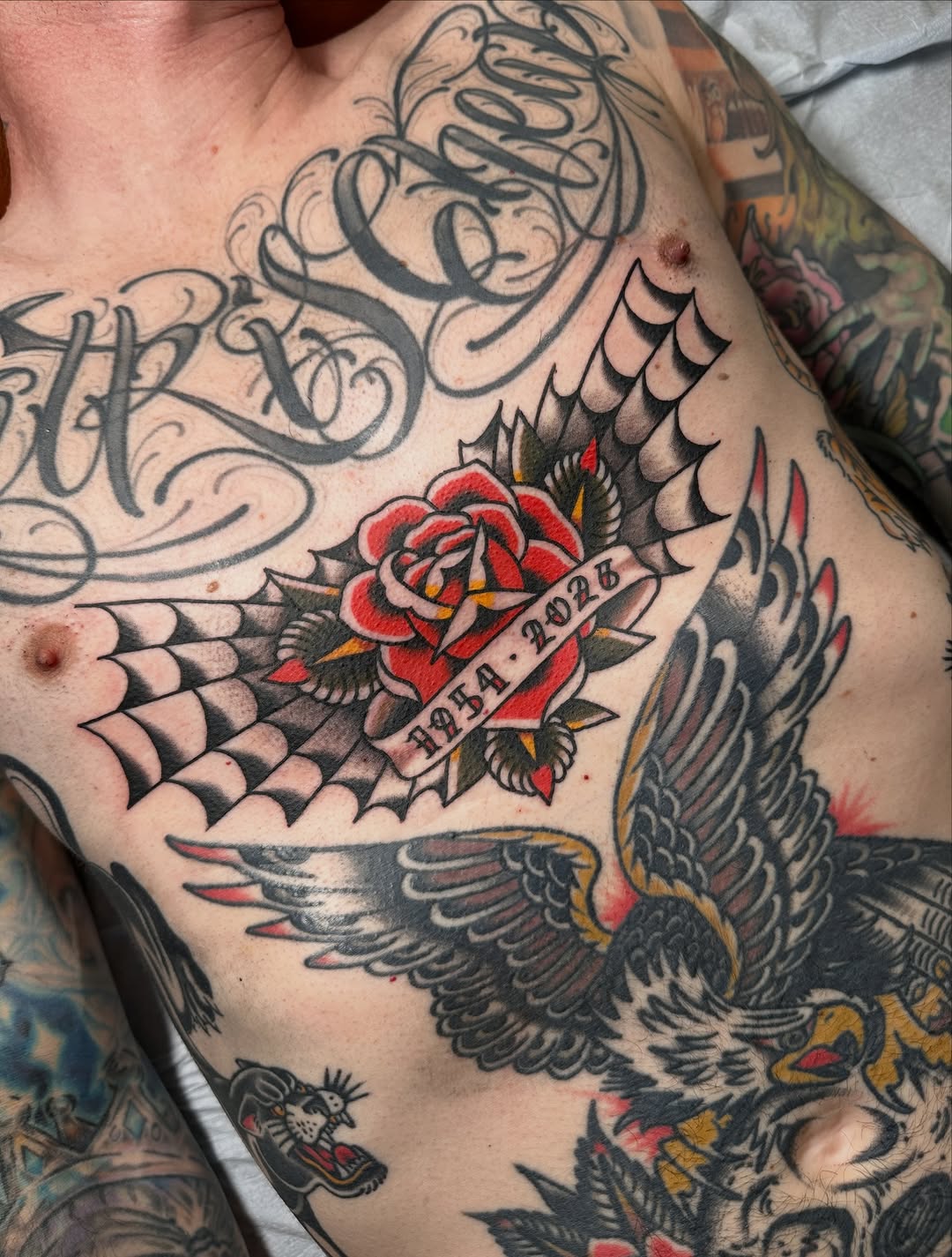 Red rose with banner containing dates, flanked by black and grey wings on chest, surrounded by ornamental scrollwork.