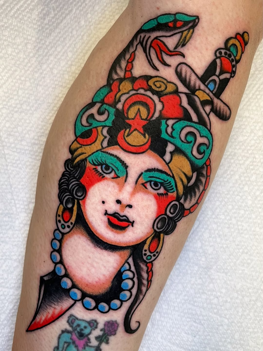 A colorful female head with red and teal face paint, ornate headdress with celestial symbols, and blue beaded necklace on the forearm.