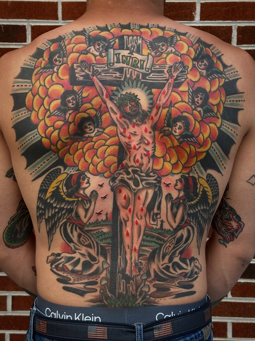 Large-scale back piece featuring a crucifix with a suffering Christ figure surrounded by ornate floral mandala and cherub imagery in bold traditional colors.