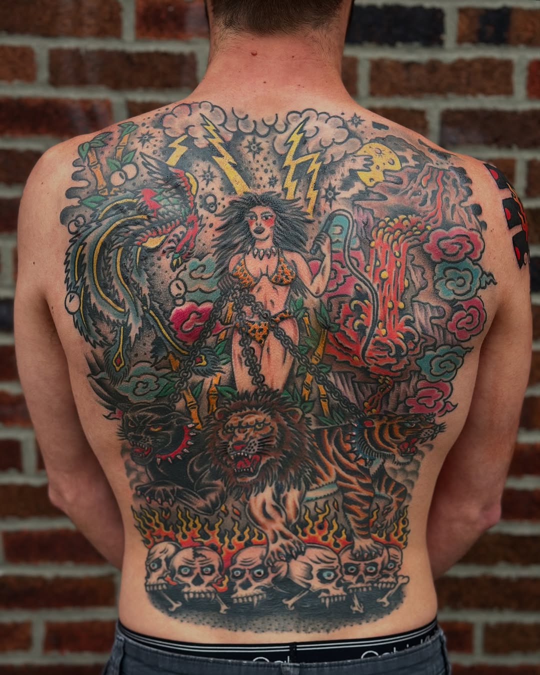 Full back piece featuring a winged female figure with lightning bolts, surrounded by tigers, roses, flames, and skulls in bold traditional American style.