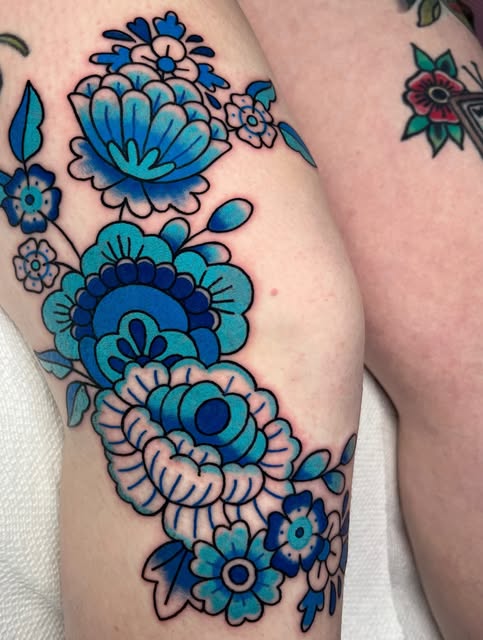 Vibrant blue and turquoise floral tattoo with layered blooms and leaves on the upper arm, featuring dark blue outlines and pink accents.