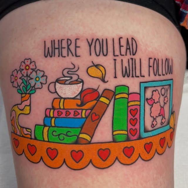 Colorful thigh tattoo featuring stacked books, flowers, teacup, watering can, and framed poodle with text 'WHERE YOU LEAD I WILL FOLLOW' and heart border.