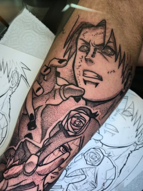 Anime character tattoo on forearm featuring expressive face with spiky hair, detailed eyes, and roses, surrounded by sketch designs on paper.