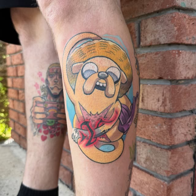 Jake the Dog from Adventure Time tattooed on a calf, wearing an orange hat and pink collar, with a blue outline and yellow/orange coloring against brick background.