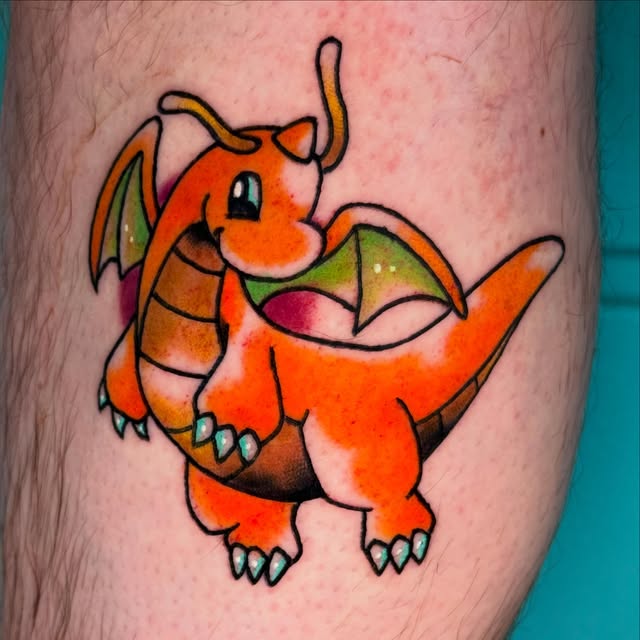Dragonite Pokémon character tattoo on leg featuring orange body, green wings, yellow horns, and pink mouth with black outlines.