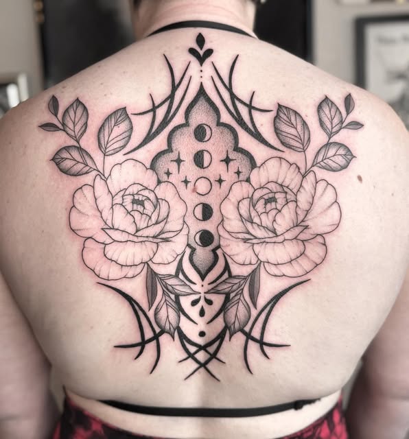 Symmetrical back tattoo featuring two peonies flanking a central cross with moon phases, botanical elements, and ornamental line work in black ink.