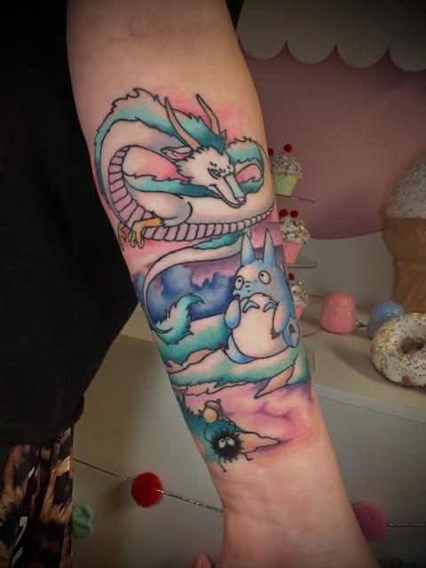 Studio Ghibli characters including a dragon and Totoro tattooed on an arm in turquoise, pink, and blue watercolor palette with decorative elements.