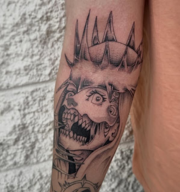 Manga-style character with spiky crown, wide grin showing sharp teeth, and mechanical side panel, tattooed on forearm in black and grey ink.