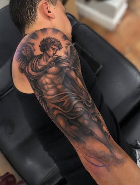 Winged male angel figure with muscular torso and halo tattoo covering the upper arm and shoulder in brown and black tones.