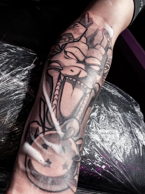 Cartoon dragon head tattoo with exaggerated features and open mouth on forearm, rendered in black and grey ink.