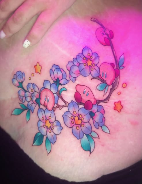 Kirby characters surrounded by purple and blue flowers with yellow centers, red stars, and teal leaves tattooed on shoulder