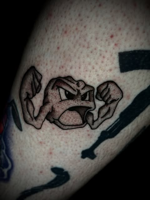 Pokémon Geodude character tattoo on upper arm, depicted with angry expression and clenched fists in black and grey ink.
