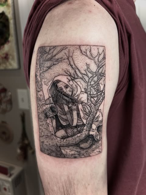 Detailed black and grey tattoo on upper arm depicting a cloaked figure among twisted branches and vines in a rectangular frame.