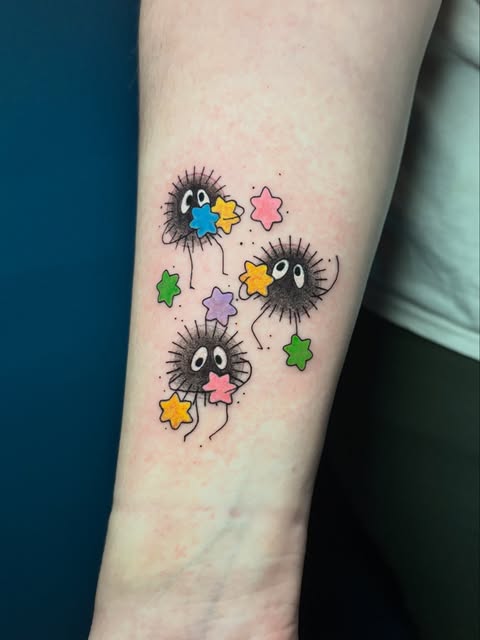 Three Totoro characters from Studio Ghibli with colorful stars scattered around them on an inner forearm, featuring blue, green, pink, and yellow accents.