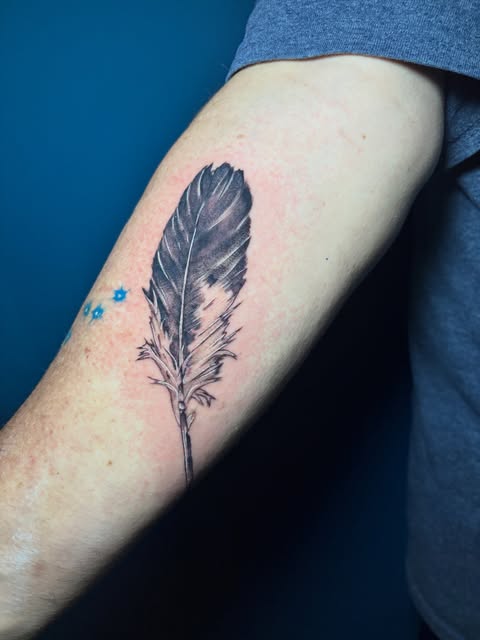 Black feather tattoo on inner forearm with small blue dots near the upper arm, against blue background.