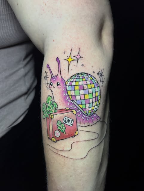 Pink sluglike creature with purple spots next to a colorful disco ball and kale plant on upper arm, surrounded by stars and sparkles.