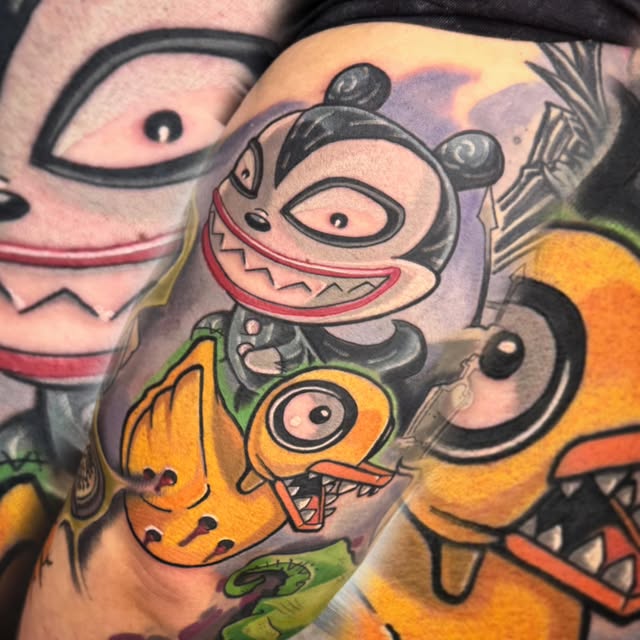 Colorful tattoo on arm featuring cartoon characters with large eyes and exaggerated grins in yellow, orange, pink, and purple tones with black outlines.