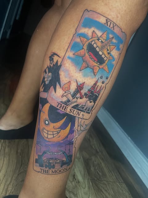 The Sun and The Moon tarot cards featuring grinning celestial characters tattooed on a person's calf in blue, orange, and purple colors.