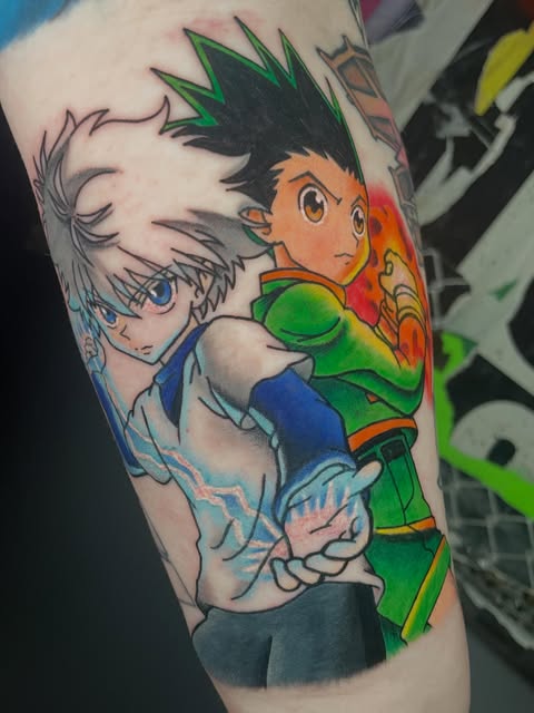 Hunter x Hunter characters Gon and Killua tattooed on an arm, featuring Gon in green and orange outfit with spiky black hair and Killua in blue with white spiky hair.