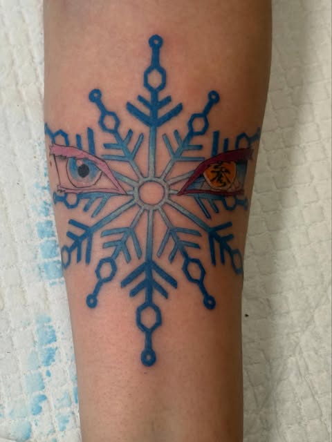 Blue snowflake tattoo on forearm with two colored eyes (one pink, one yellow/orange) intersecting horizontally through the center.