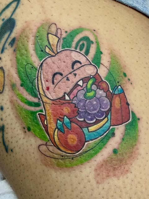 Kirby from Nintendo wearing chef's hat and apron, holding grapes and food items, tattooed on shoulder with green background, brown, purple, and blue accents.