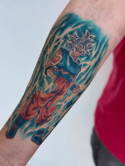 Goku from Dragon Ball Z tattoo on forearm in red gi and spiky hair, surrounded by blue energy effects and motion lines.