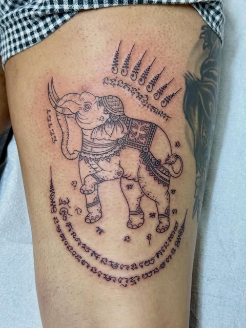Ornate elephant with decorative patterns and Thai script tattoo on thigh, featuring intricate line work in black ink.