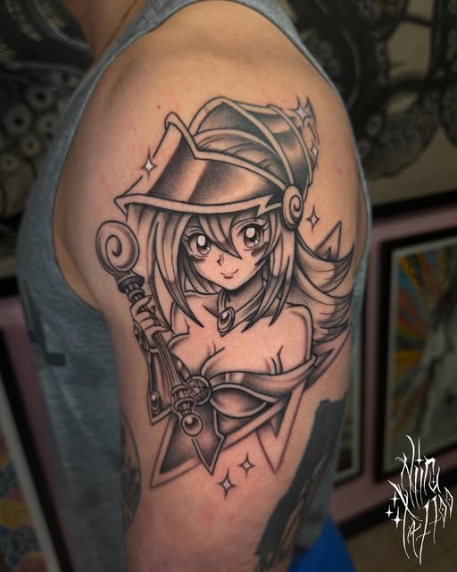 Anime girl character with long hair wearing a witch hat and holding a staff, tattooed on upper arm in black and grey ink with decorative stars.