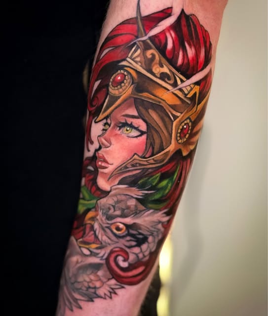 Elaborate tattoo on forearm featuring a woman with red feathered headdress, gold ornamental mask, and a dragon below, rendered in vivid reds, golds, greens, and skin tones.