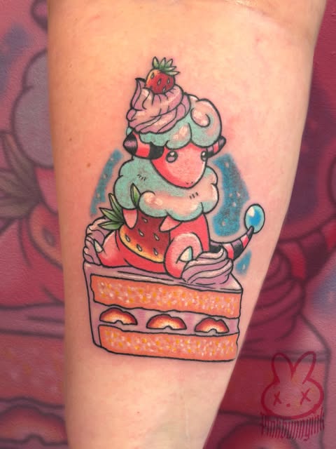 Colorful tattoo on upper arm featuring a whimsical strawberry cake character with pink frosting, green leaves, and a strawberry on top, stacked on an orange cake base.