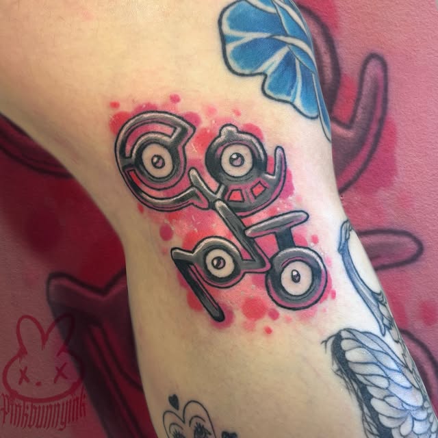 Pokémon Unown characters tattoo on upper arm with pink watercolor background and blue butterfly designs on the sides.
