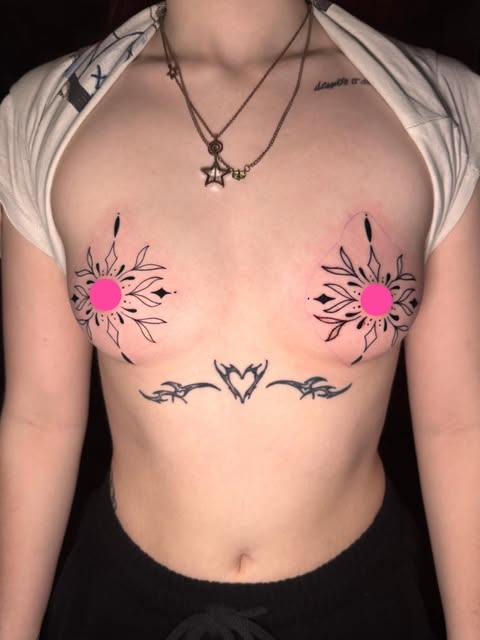 Chest tattoo featuring two pink-centered sun/flower designs on pectorals and a tribal heart design below sternum on shirtless person wearing necklace.
