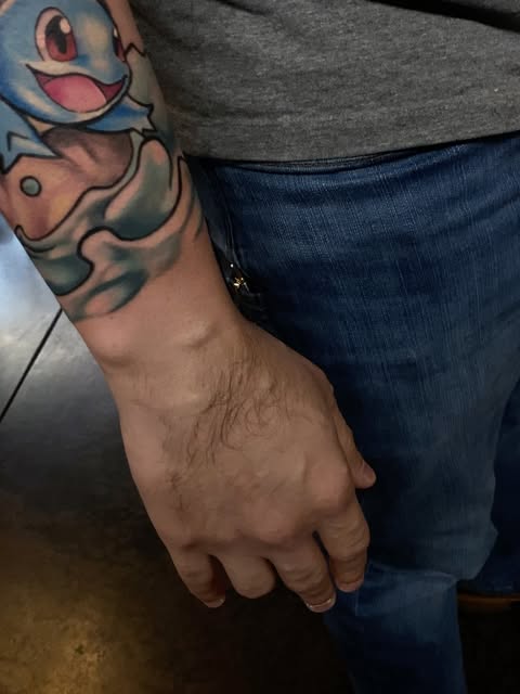 Pokémon Squirtle character tattoo on forearm featuring blue shell, pink belly, and red eyes with details on upper arm.