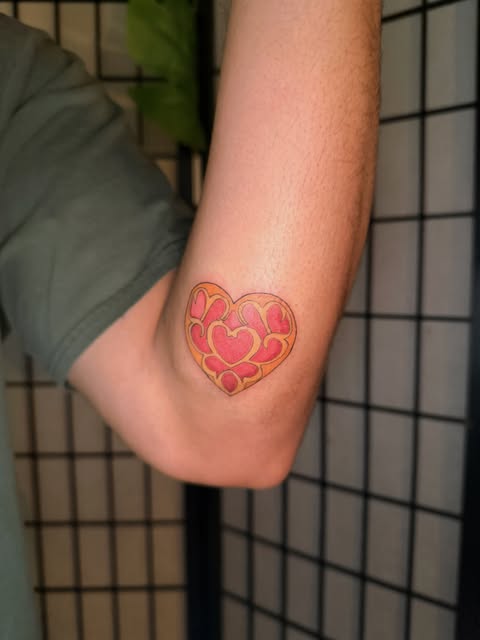 Heart-shaped tattoo composed of smaller pink and red hearts with gold/yellow outlines on the inner elbow of a pale-skinned arm against a dark grid background.