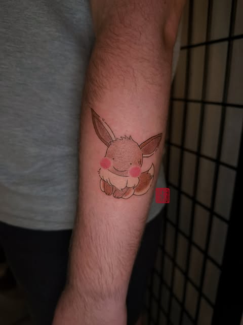 Eevee from Pokémon tattooed on an inner forearm with pink cheek blush marks and brown outline details.