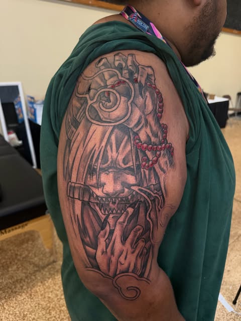 Aztec or Mayan deity tattoo on upper arm featuring an ornate mask with spiral eye, feathered headdress, and skeletal jaw in red and black ink.