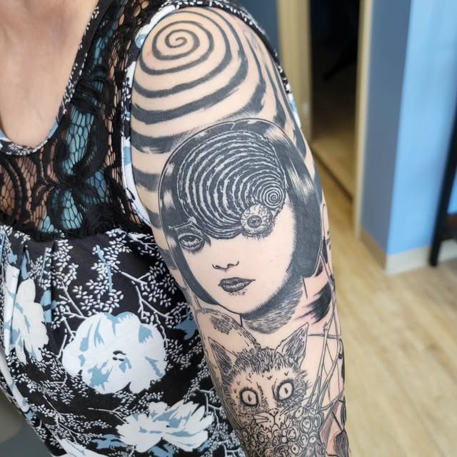 Black and grey tattoo on upper arm featuring a woman's face with spiral patterns covering her eyes, surrounded by geometric spirals and smaller illustrated elements including an owl.