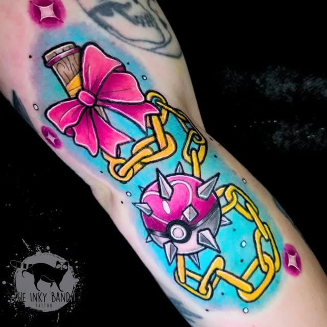 Colorful tattoo on forearm featuring a pink bow, spiked heart, golden chain links, and a brush against a blue watercolor background with sparkles.