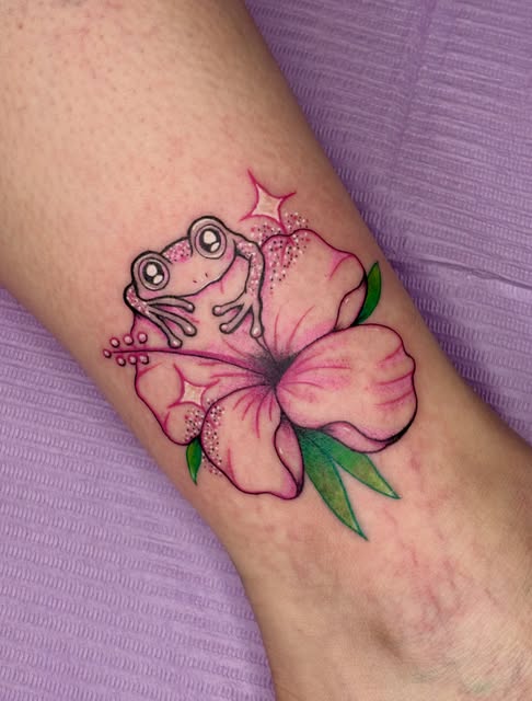 A pink hibiscus flower with a cartoon frog sitting on the petals, green leaves, and a pink star, tattooed on an arm.