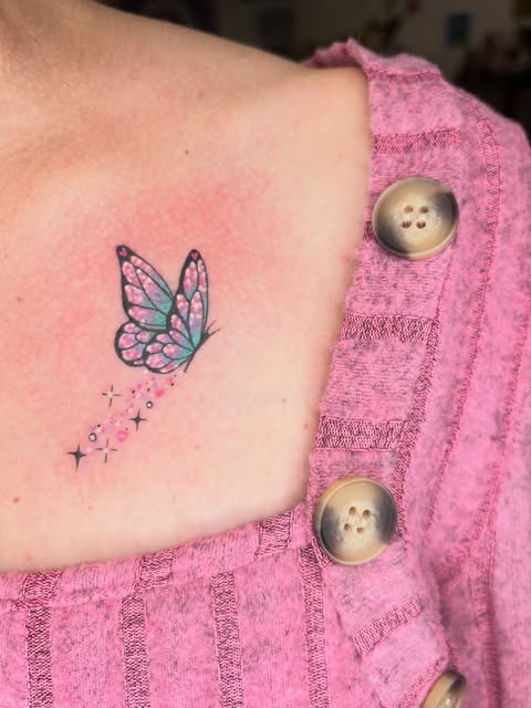 Butterfly tattoo with pink and teal wings and trailing sparkles on inner arm, worn with pink cardigan.