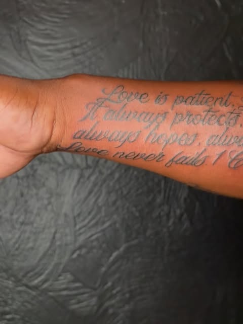 Inspirational quote tattoo on forearm reading 'Love is patient, I always protects, always trusts, always hopes, always perseveres, Love never fails' in blue-grey script.