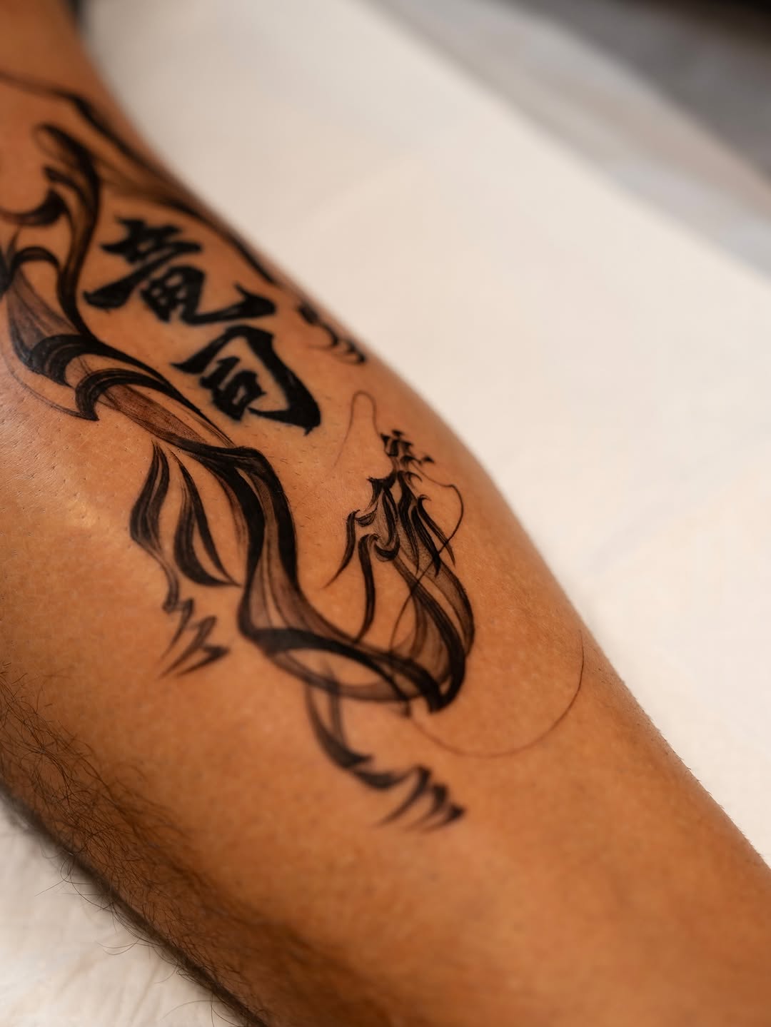 Ornate black floral and script tattoo on upper arm featuring detailed petals, flowing lines, and Asian characters with decorative frame elements.
