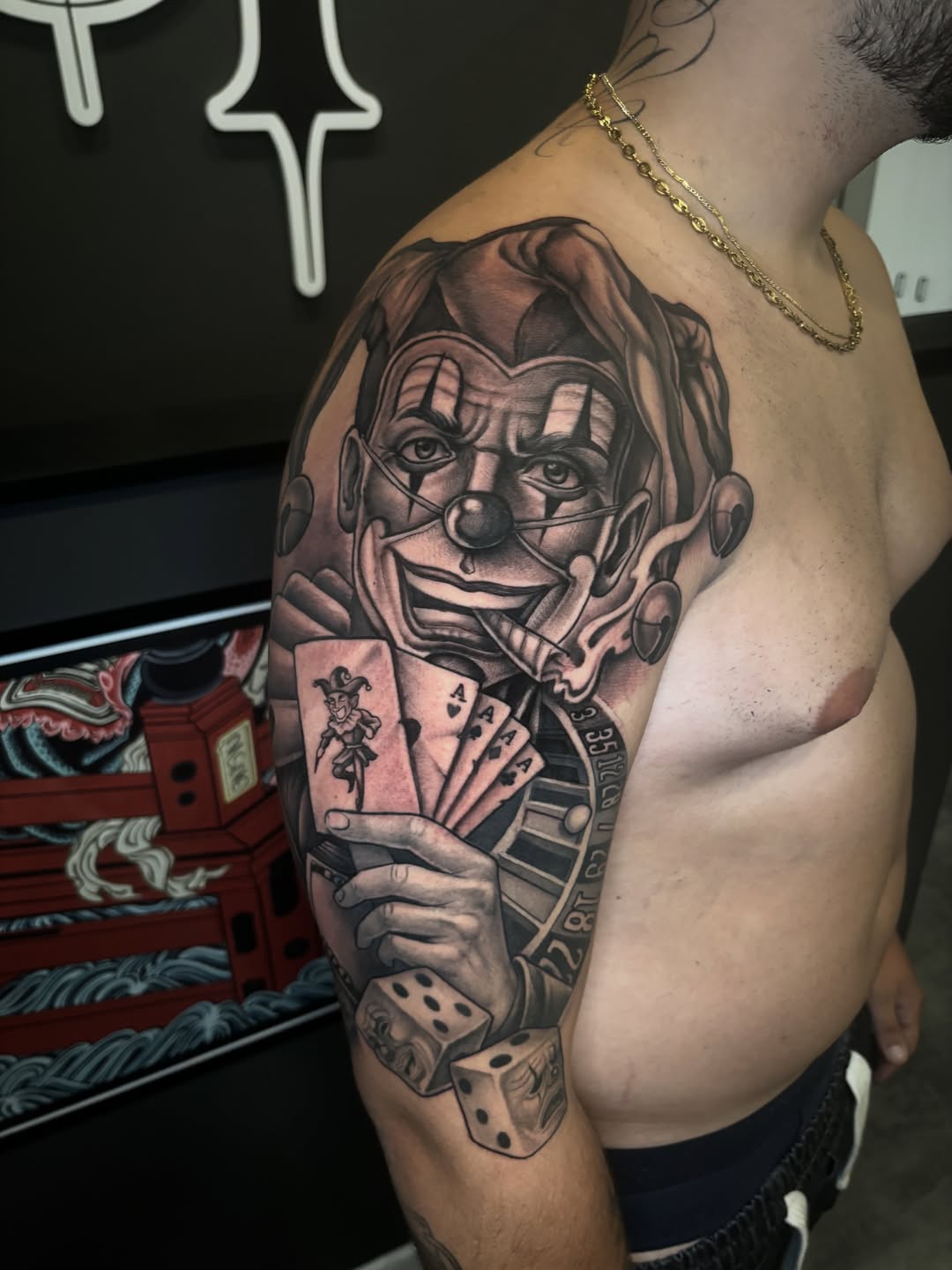 Black and grey joker clown face tattoo with playing cards and dice on upper arm and shoulder.