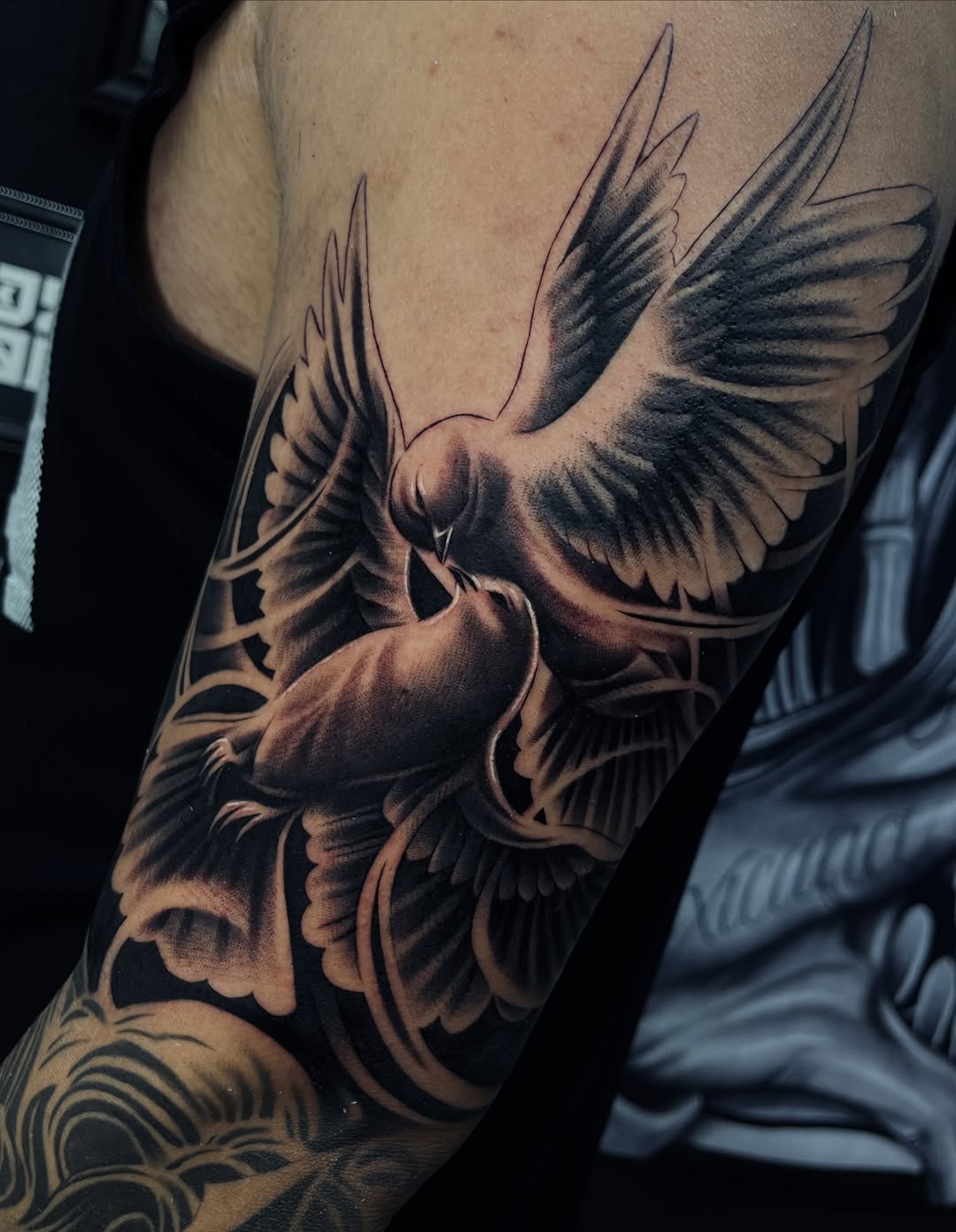 Black and grey tattoo of two doves beak to beak with spread wings, shaded in dark tones, covering the upper arm and shoulder.